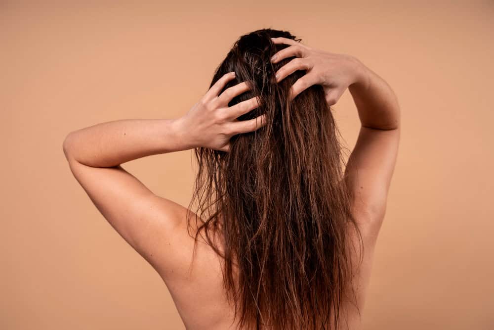 A staged photo of a woman with long dark hair facing a neutral backdrop. Her hair is damp and she is massaging her scalp.