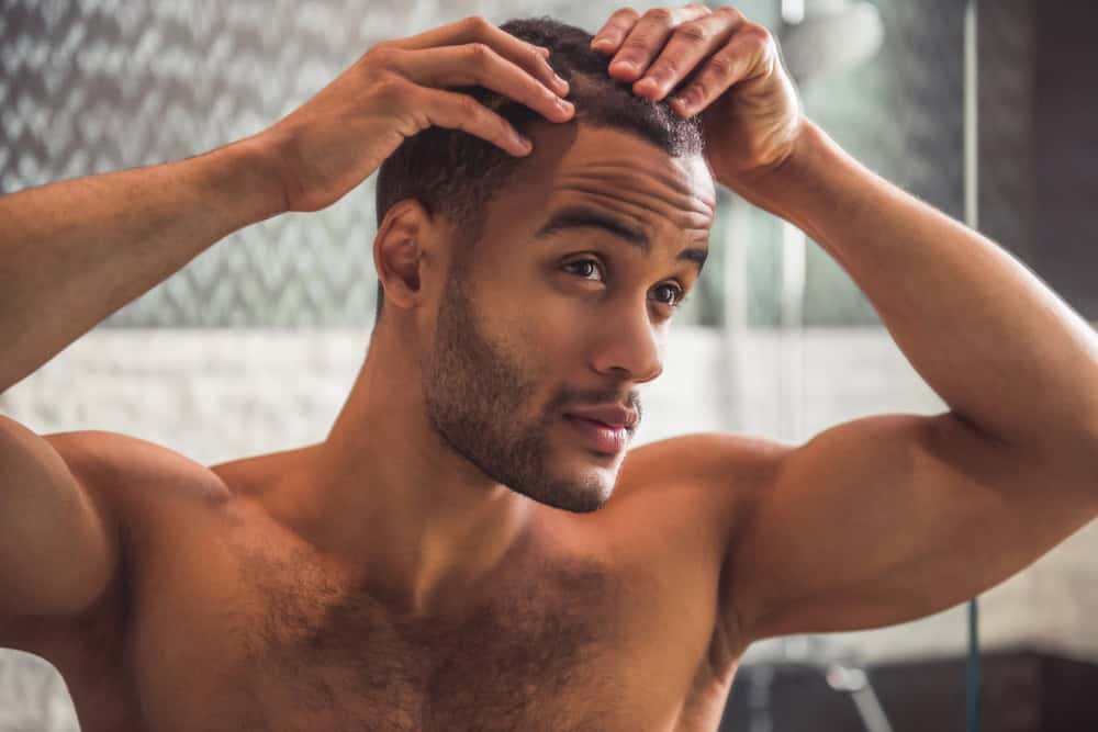 A shirtless man stands in his bathroom looking in the mirror to check his hairline
