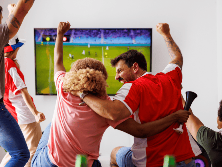 A family watch the football at home, celebrating at a goal. The angle shows the back of them celebrating and the football on the television