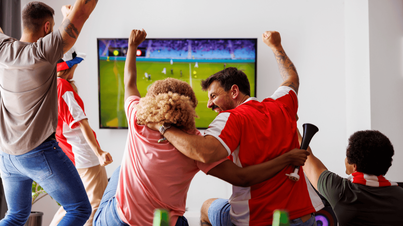A family watch the football at home, celebrating at a goal. The angle shows the back of them celebrating and the football on the television