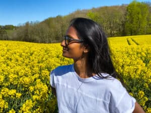 A beautiful woman with dark hair stands in a field of yellow flowers with sunglasses looking away