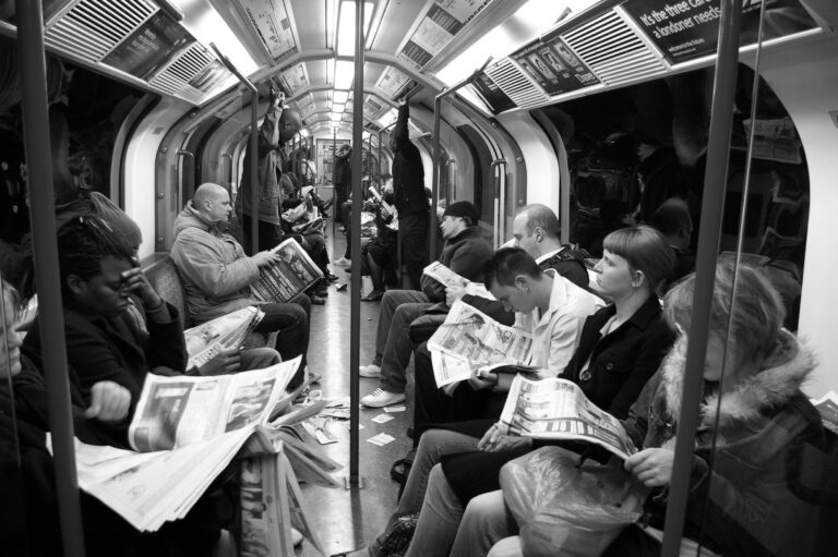 passengers inside a London Underground tube carriage, with multiple individuals reading newspapers, illustrating how unspoken social norms and conformity dictate acceptable public behaviour