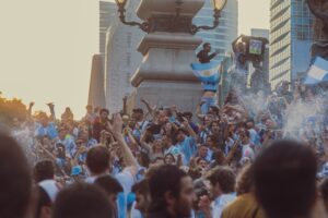 dense crowd of ecstatic football fans celebrating outdoors in a city plaza, with individuals wearing light blue and white striped jerseys, waving flags featuring a central sun emblem, holding up a replica gold trophy, and celebrating amidst a spray of water and confetti with modern skyscrapers in the background.