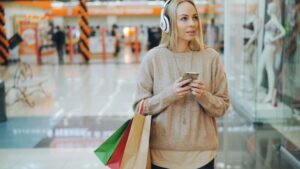 A shopper wearing headphones and using a smartphone in a retail store, illustrating how consumers integrate digital technology into physical shopping environments