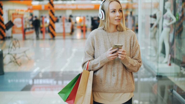 A shopper wearing headphones and using a smartphone in a retail store, illustrating how consumers integrate digital technology into physical shopping environments