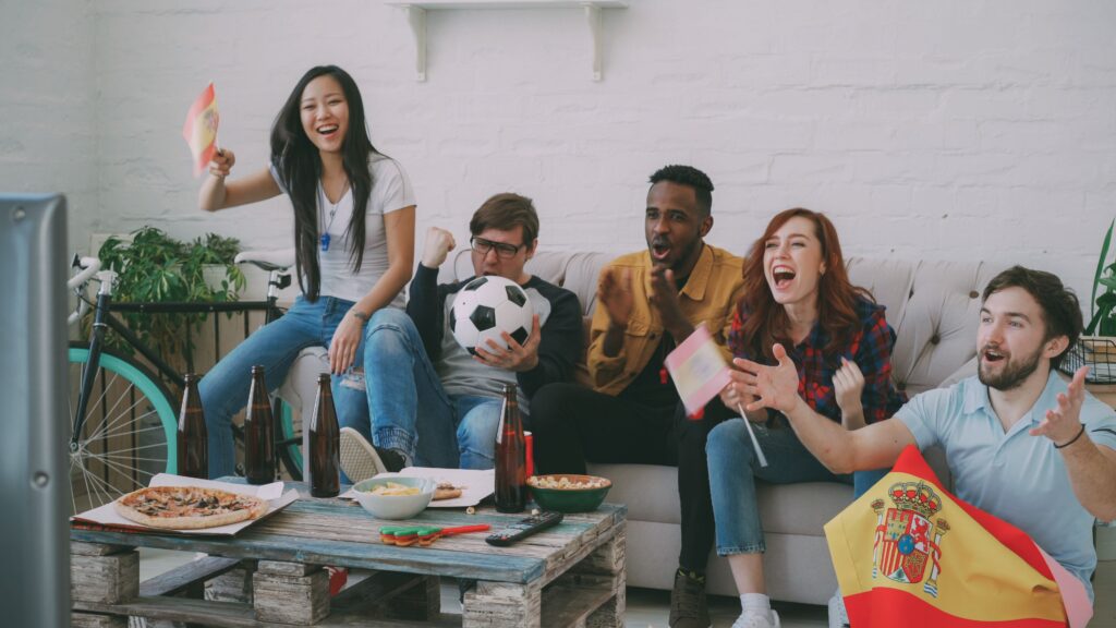 A group of friends on a sofa cheering while watching a football match on television, with individuals holding small Spanish flags and a football, in front of a coffee table layout with a pizza, snacks, and drinks.