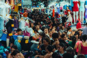 A bustling, densely crowded clothing market filled with individuals making purchases, illustrating a complex retail environment where shopper buying habits and end-consumer choices diverge