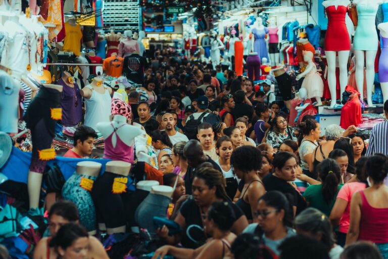 A bustling, densely crowded clothing market filled with individuals making purchases, illustrating a complex retail environment where shopper buying habits and end-consumer choices diverge